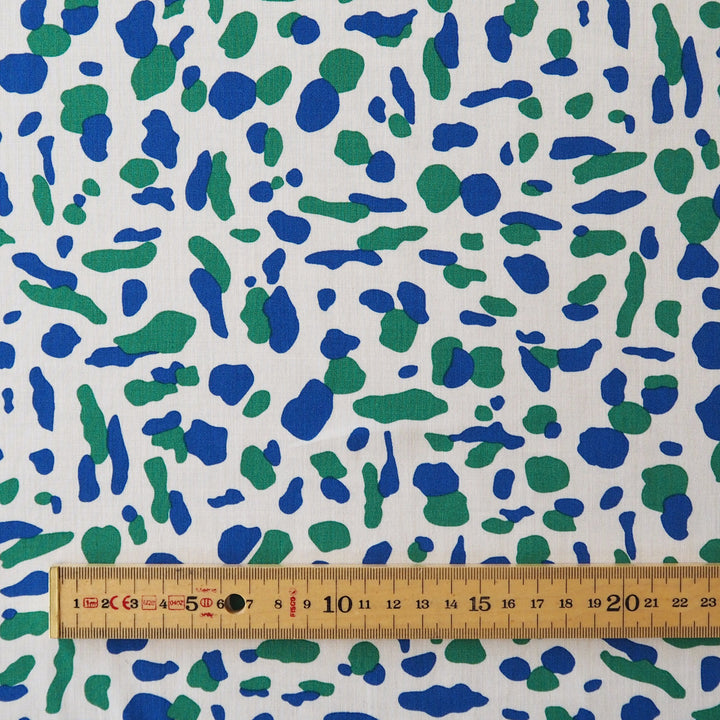 White fabric with an abstract blue and green spotted pattern, shown with a wooden ruler at the bottom for scale.