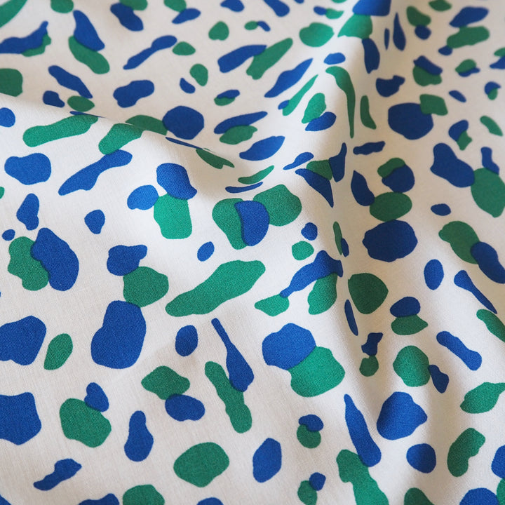 A close-up of a fabric with an abstract organic spot pattern in blue and green on a warm off-white background.