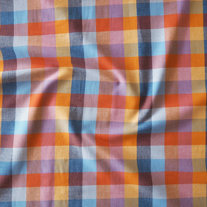 A close-up of a plaid fabric with vertical and horizontal stripes in orange, blue, yellow, purple, and white, showing a soft draped texture.