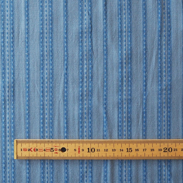 A close-up of light blue striped fabric with vertical stitched lines, shown with a wooden ruler at the bottom for scale.