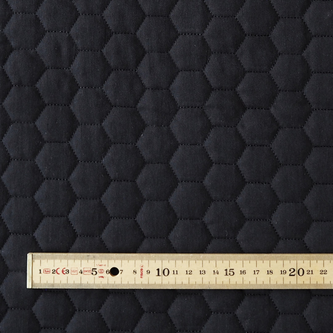 Close-up of a dark charcoal gray fabric with a stitched hexagonal quilted pattern, with a wooden ruler at the bottom showing the scale of the hexagons.