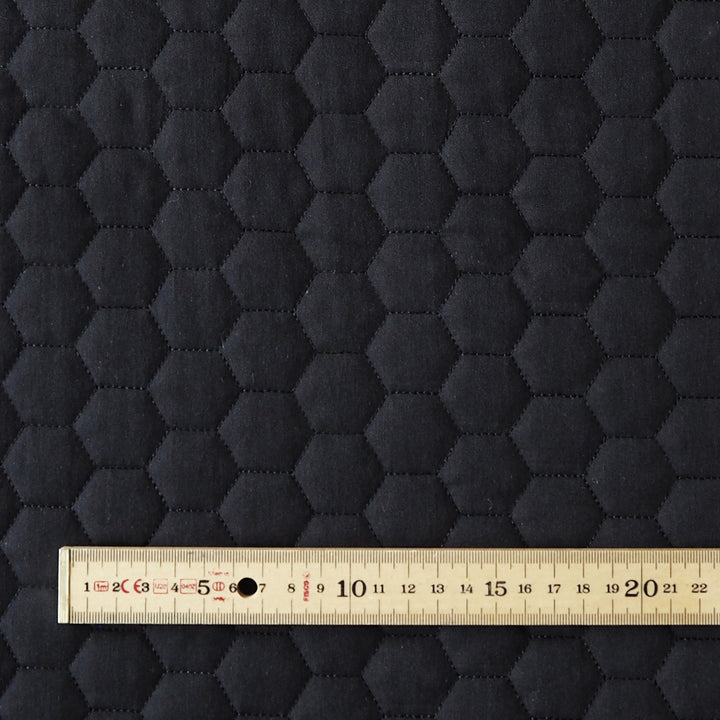 Close-up of a dark charcoal gray fabric with a stitched hexagonal quilted pattern, with a wooden ruler at the bottom showing the scale of the hexagons.