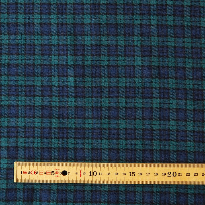 A close-up of tartan fabric in dark blue and teal green with a wooden ruler along the bottom edge for scale.