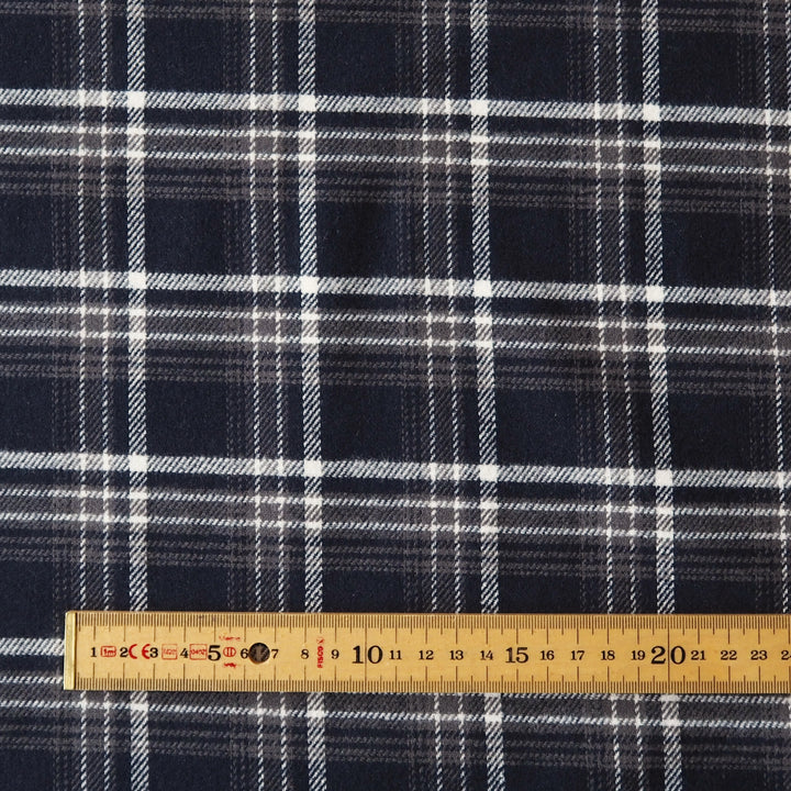A close-up of navy blue plaid fabric with white and gray intersecting stripes, shown with a wooden measuring tape ruler along the bottom edge for scale.