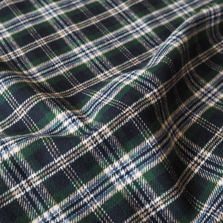 A close-up of a plaid fabric with a classic tartan pattern in dark green, navy blue, white, and thin light blue stripes, shown with soft folds.