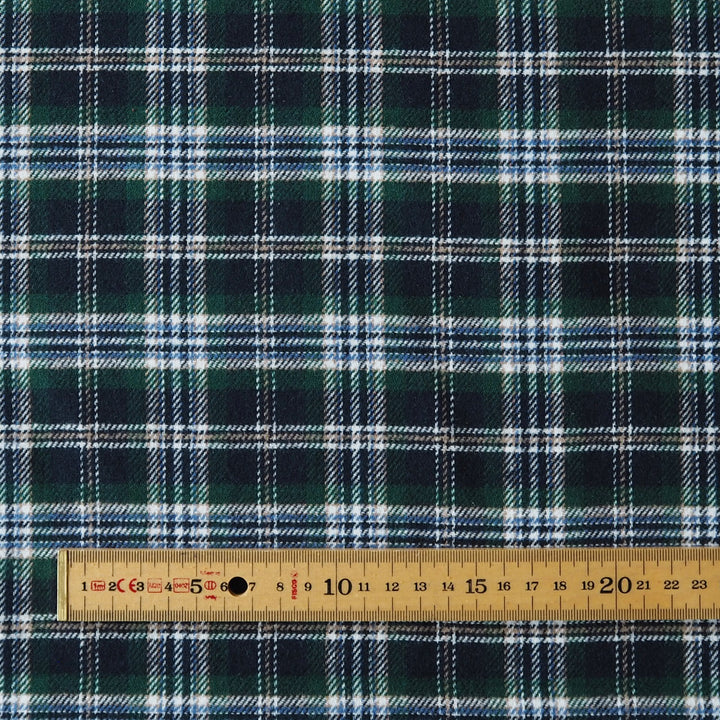 A close-up of dark green plaid fabric with white and light blue intersecting stripes, with a wooden ruler along the bottom edge showing measurement markings.