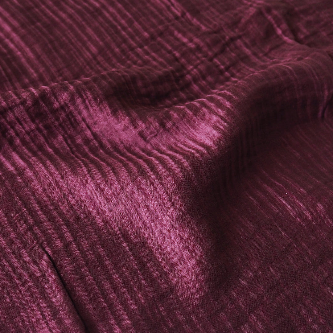 A close-up of textured fabric in a deep plum purple color, showing a crinkled or gauze-like weave with soft folds and shadows.