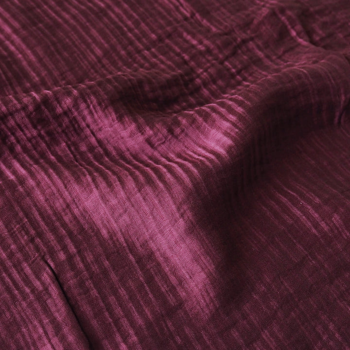 A close-up of textured fabric in a deep plum purple color, showing a crinkled or gauze-like weave with soft folds and shadows.