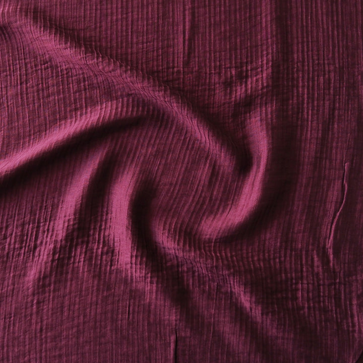 A close-up of draped crinkled gauze fabric in a deep plum purple color, showing the textured weave and soft folds.