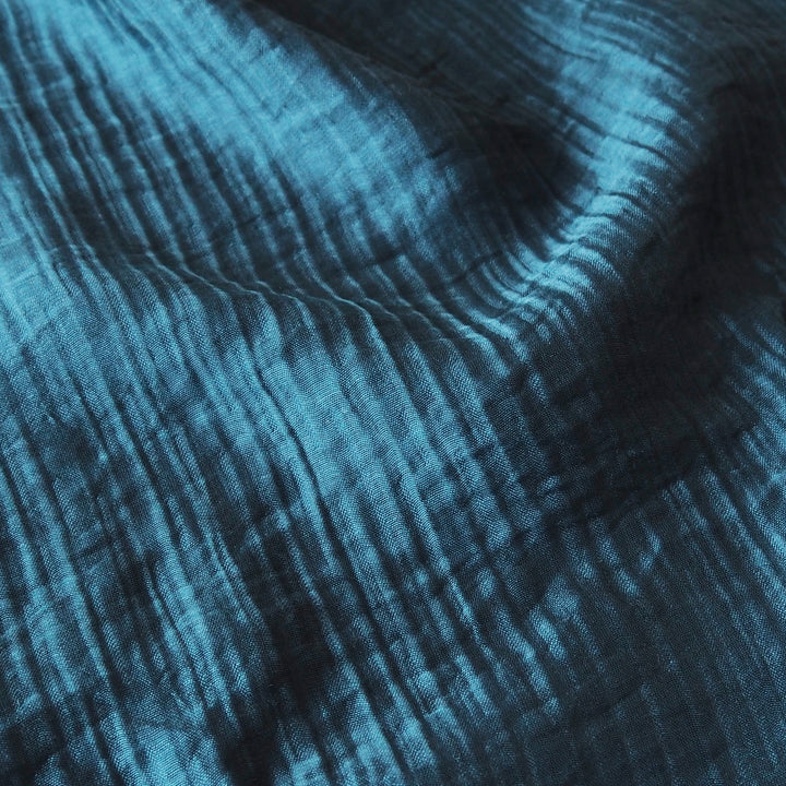 A close-up of textured blue fabric with a crinkled gauze or muslin pattern, showing folds and shadows.