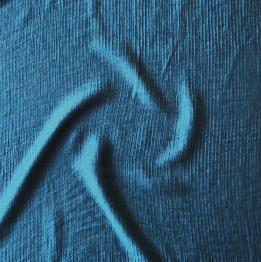 A close-up of textured blue fabric with a crinkled, gauze-like surface and soft folds.