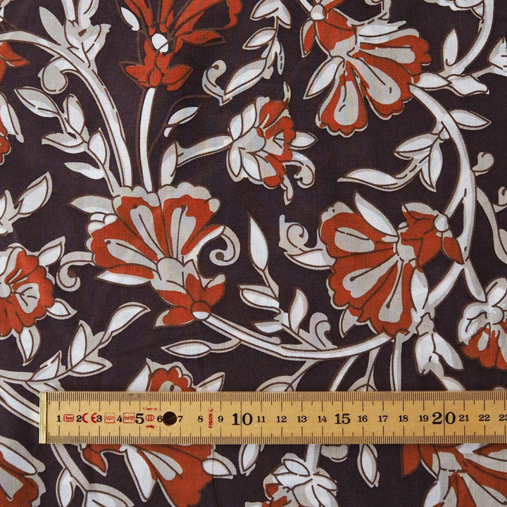A close-up of a floral fabric pattern with rust red and white flowers on a dark charcoal background, with a wooden ruler at the bottom showing the scale of the print.