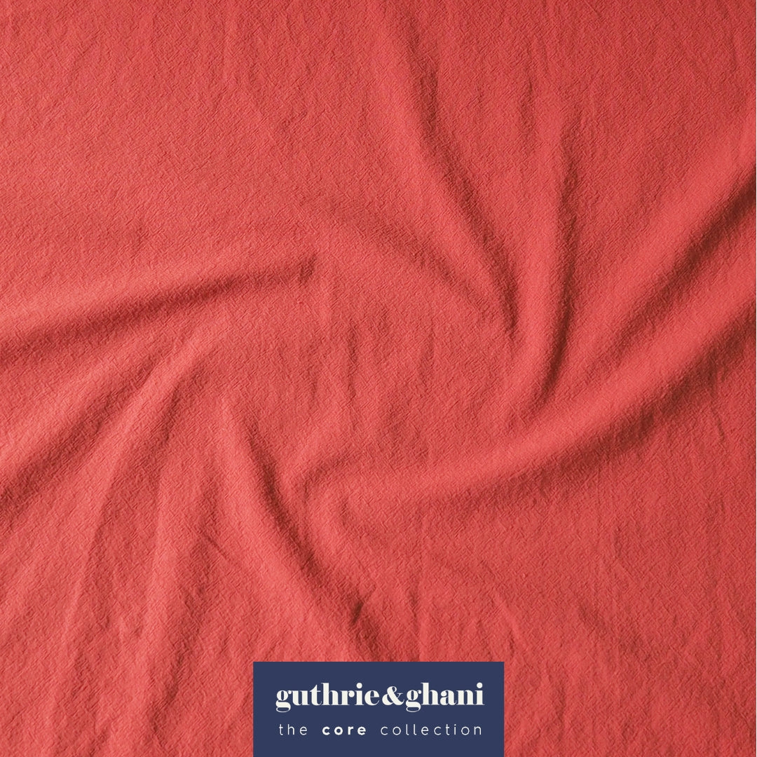A close-up of coral pink textured fabric with a swirl pattern, featuring a navy blue brand label at the bottom that reads 'guthrie&ghani the core collection'.