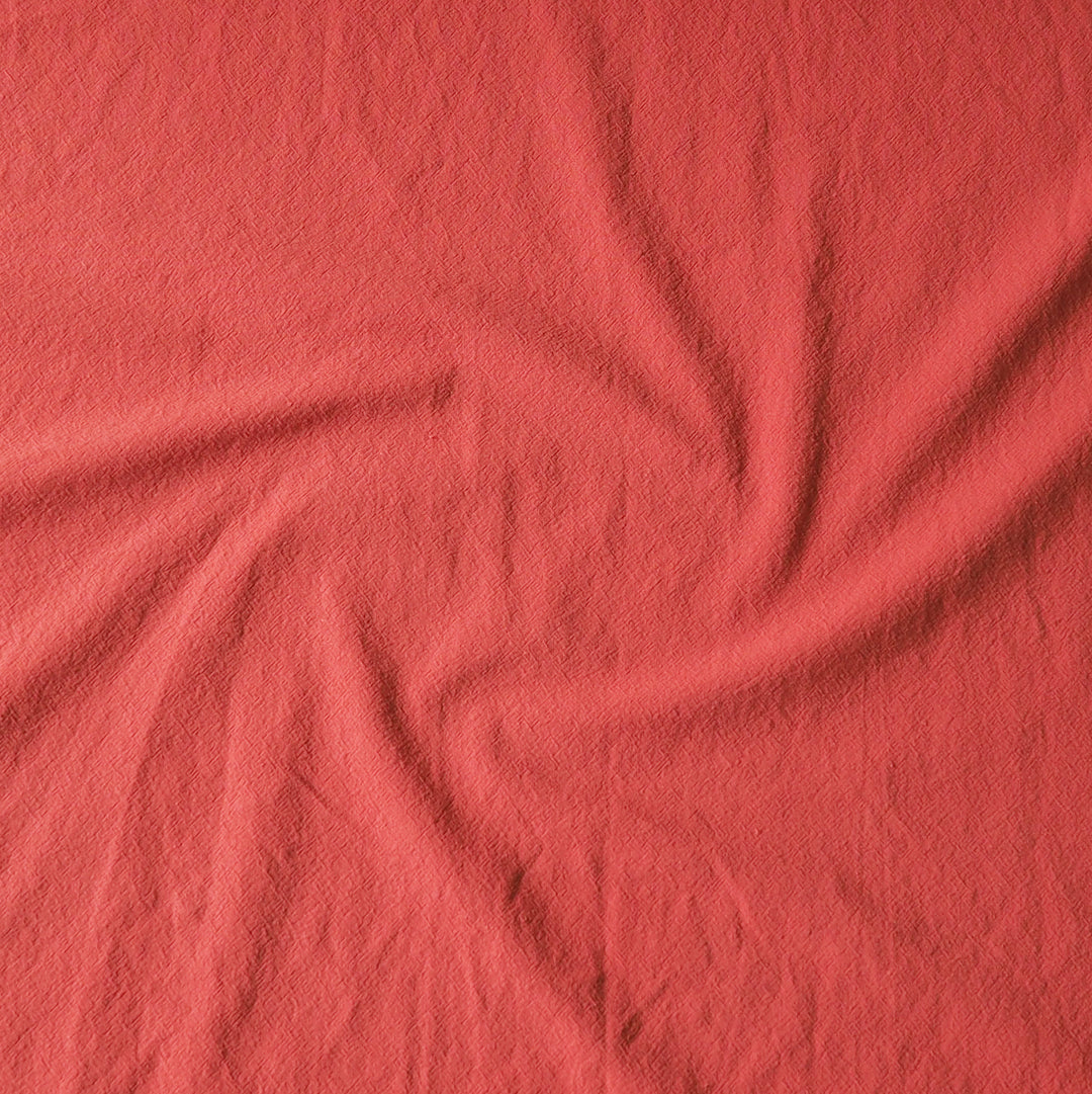 A close-up of textured fabric in a solid coral pink color, arranged with a central swirl of folds.
