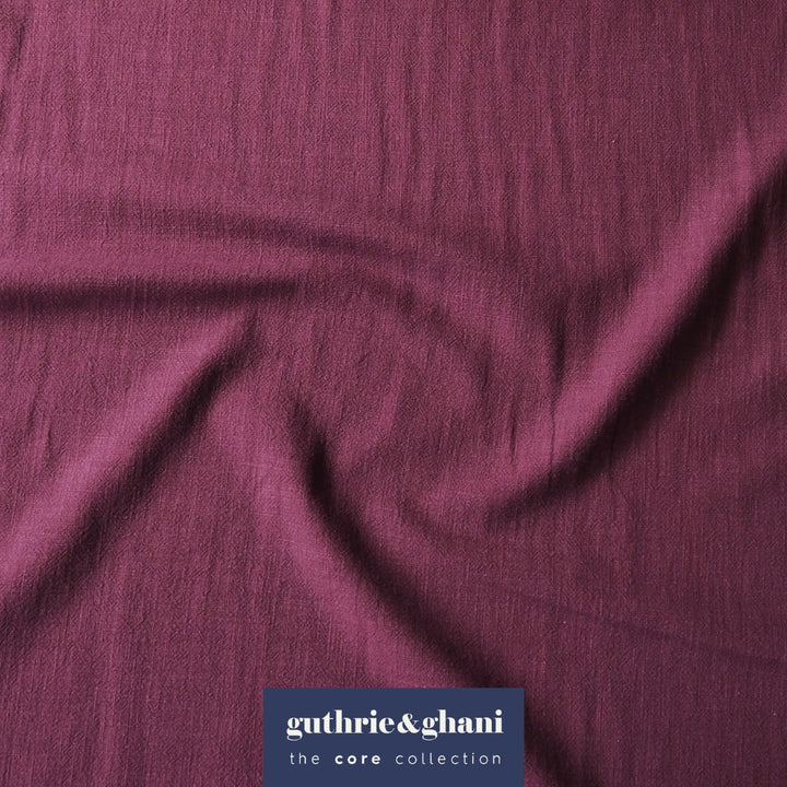 A close-up of draped mauve-purple fabric with a subtle crinkled texture, featuring a dark blue brand label at the bottom that reads 'guthrie&ghani the core collection'.