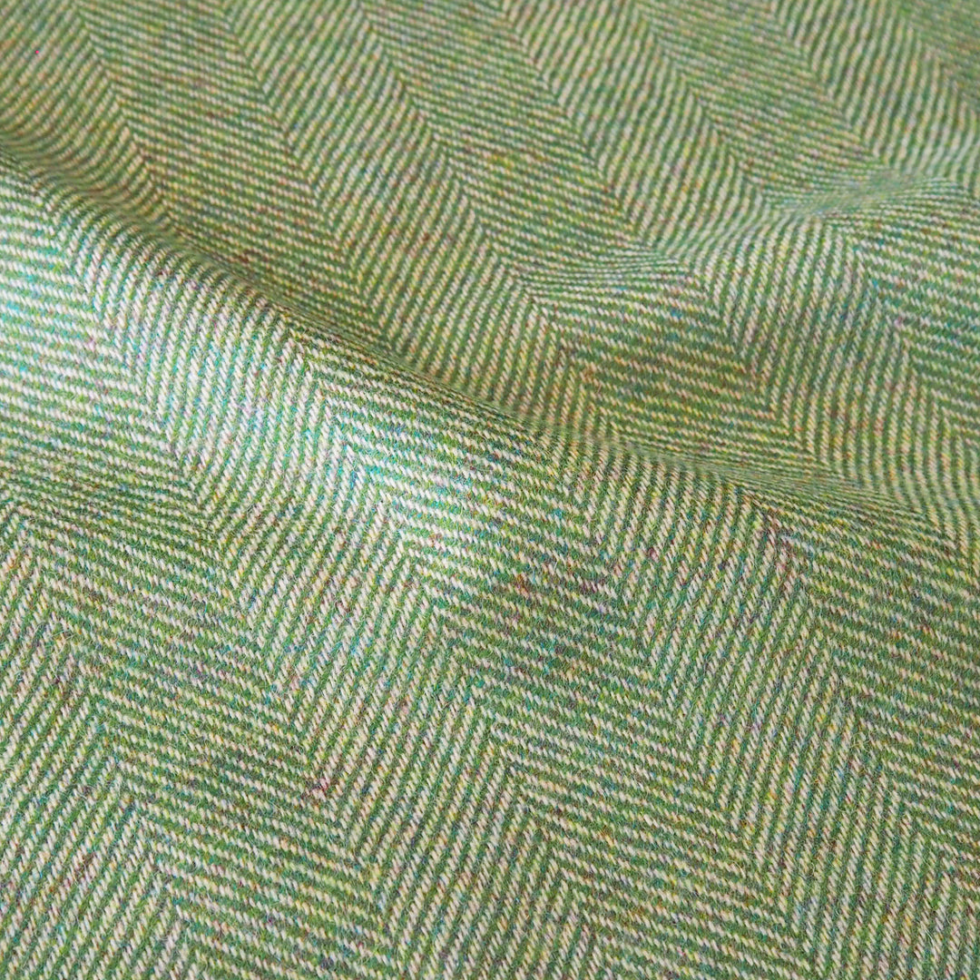 A close-up of woven fabric with a herringbone pattern in muted green, beige, and gray tones, showing the textile texture and folds.