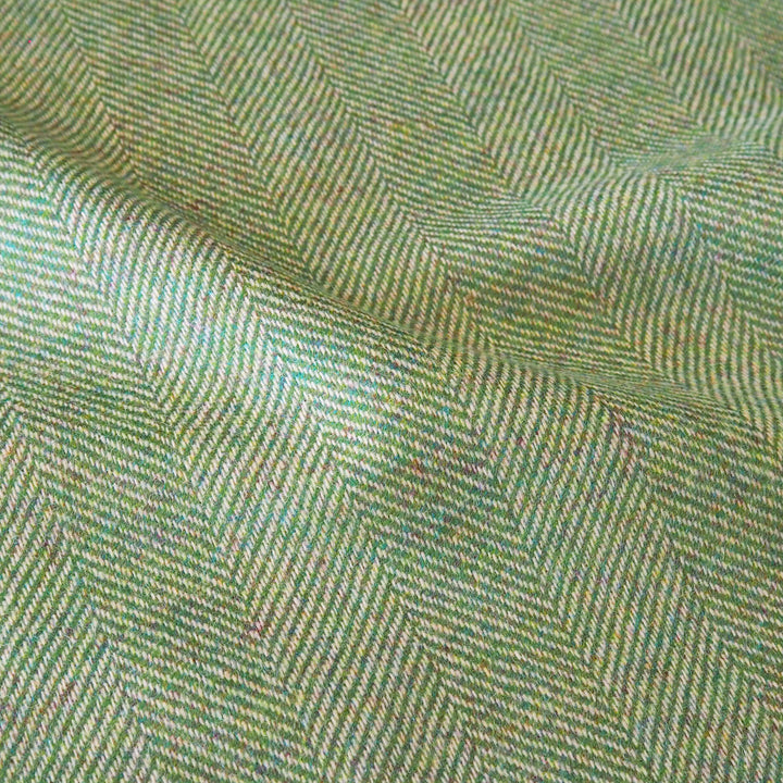 A close-up of woven fabric with a herringbone pattern in muted green, beige, and gray tones, showing the textile texture and folds.
