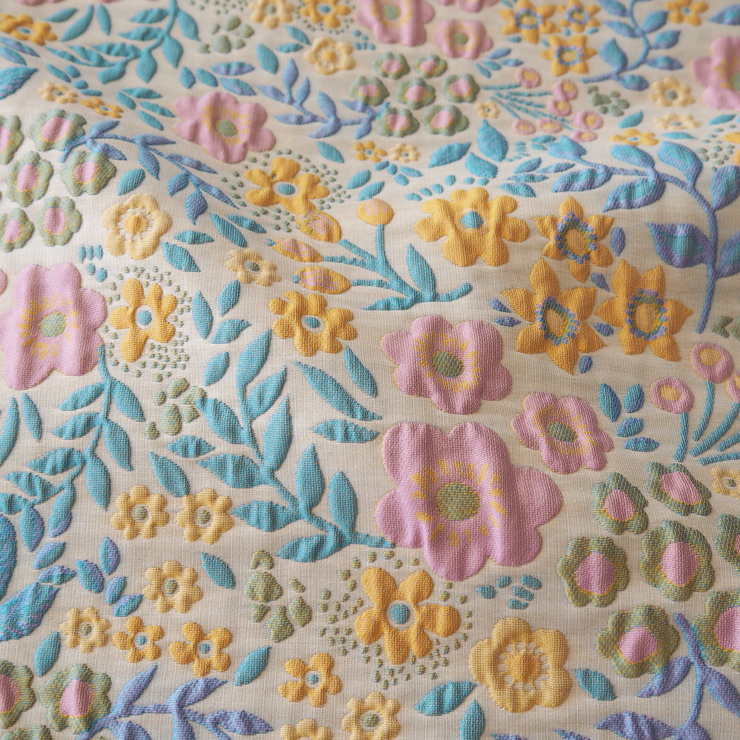 A close-up of a textured fabric featuring a colorful floral pattern with pink, yellow, and light blue embroidered flowers and leaves on a cream-colored woven background.