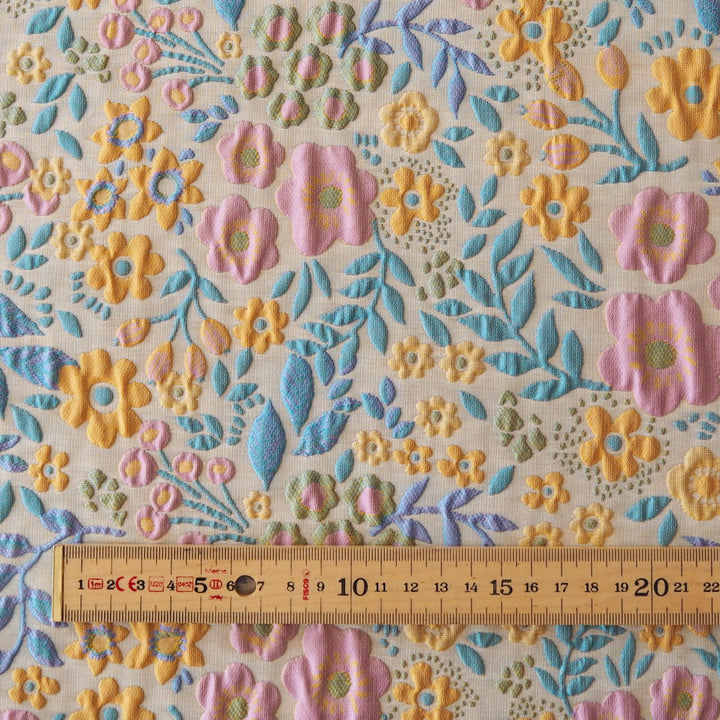 A close-up of a floral fabric pattern with embroidered or raised flowers and leaves in pink, yellow, blue, and green on a light beige background, with a wooden ruler along the bottom edge for scale.