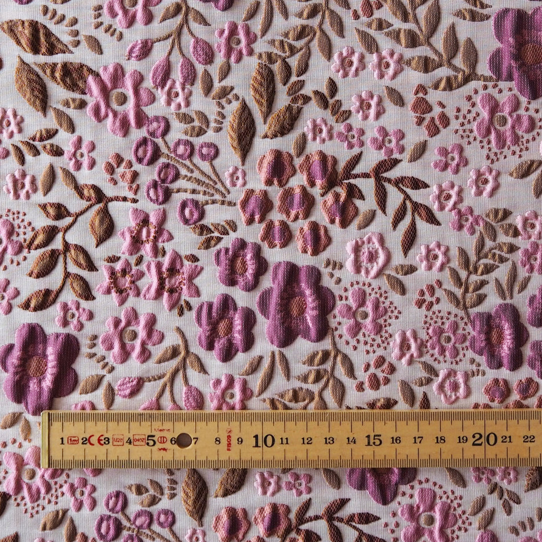 Close-up of a floral embroidered fabric with pink and mauve flowers, brown leaves, and a wooden ruler along the bottom edge for scale.