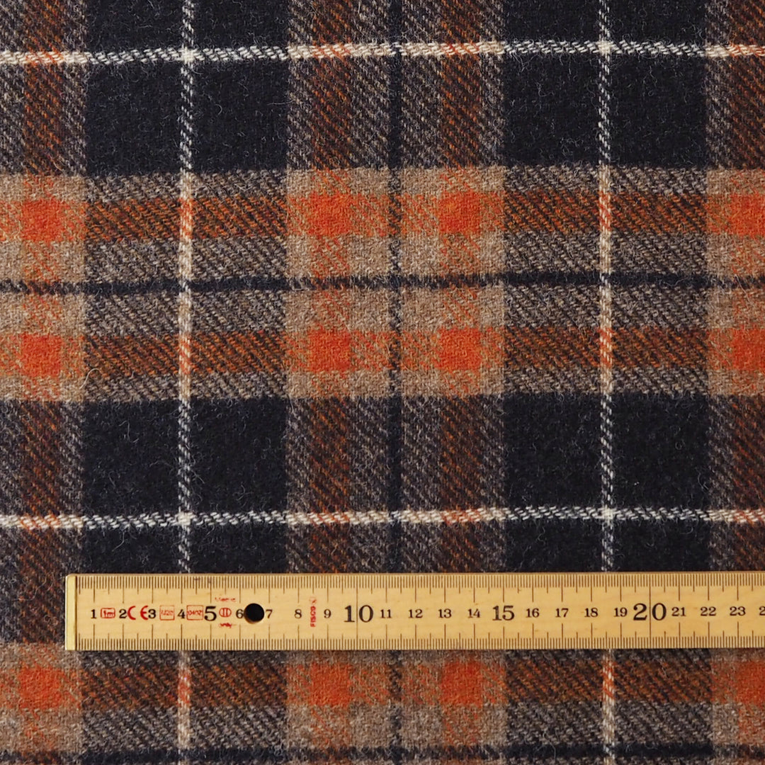 A close-up of a plaid fabric pattern with dark navy, black, rust orange, and beige stripes, shown with a wooden ruler at the bottom for scale.