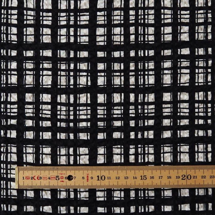 Black and white plaid fabric pattern shown close up with a wooden ruler at the bottom for scale.