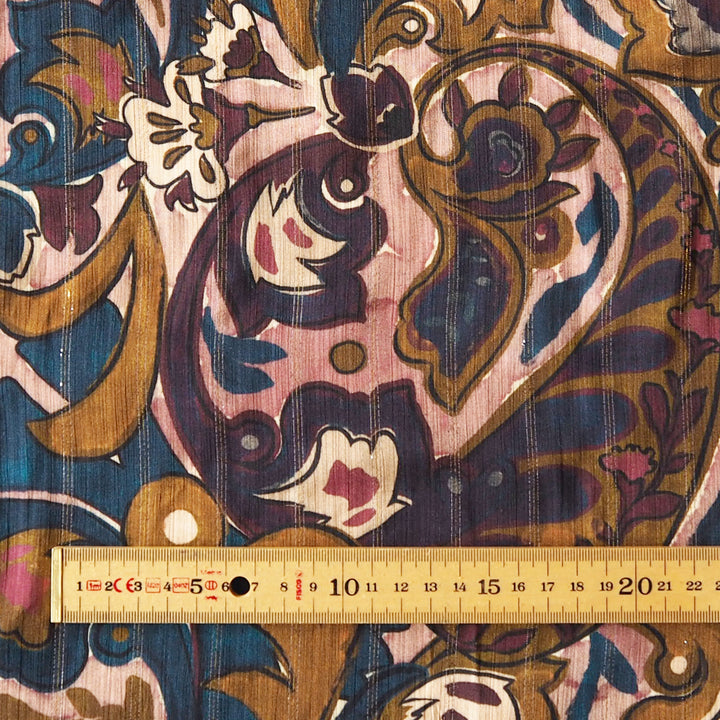 A close-up of patterned fabric with a large ornate paisley/floral design in navy, blue, pink, gold, and white, with a wooden ruler along the bottom edge for scale.