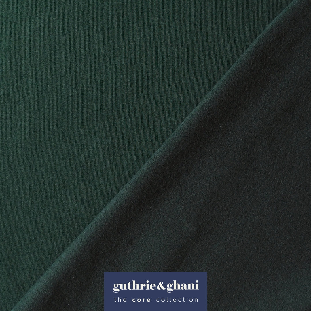 A close-up of dark forest green fabric with a diagonal fold, featuring a blue brand label at the bottom that reads 'guthrie&ghani the core collection'.