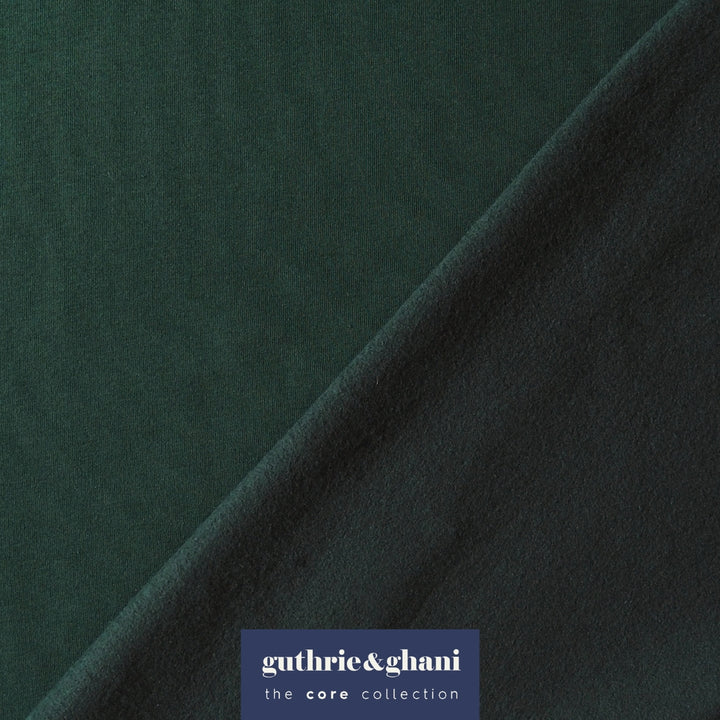 A close-up of dark forest green fabric with a diagonal fold, featuring a blue brand label at the bottom that reads 'guthrie&ghani the core collection'.