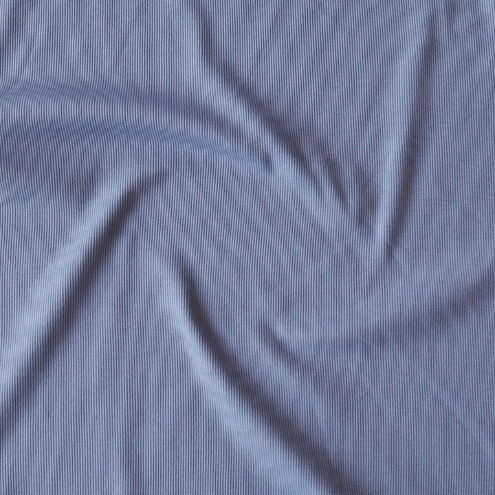 A close-up of a light periwinkle blue ribbed fabric, showing its fine vertical texture and soft folds.