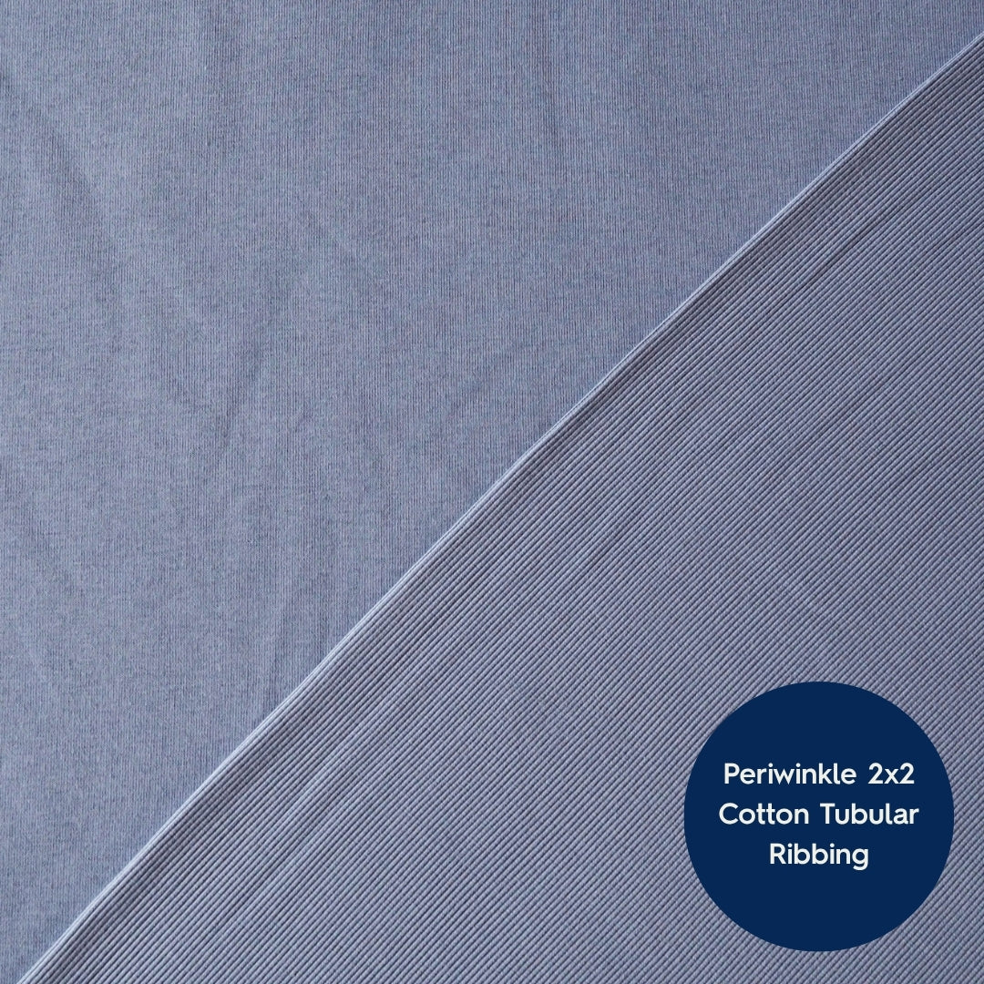 A close-up of periwinkle blue cotton tubular ribbed fabric, showing a diagonal fold line and a dark blue circular label with the text 'Periwinkle 2x2 Cotton Tubular Ribbing'.