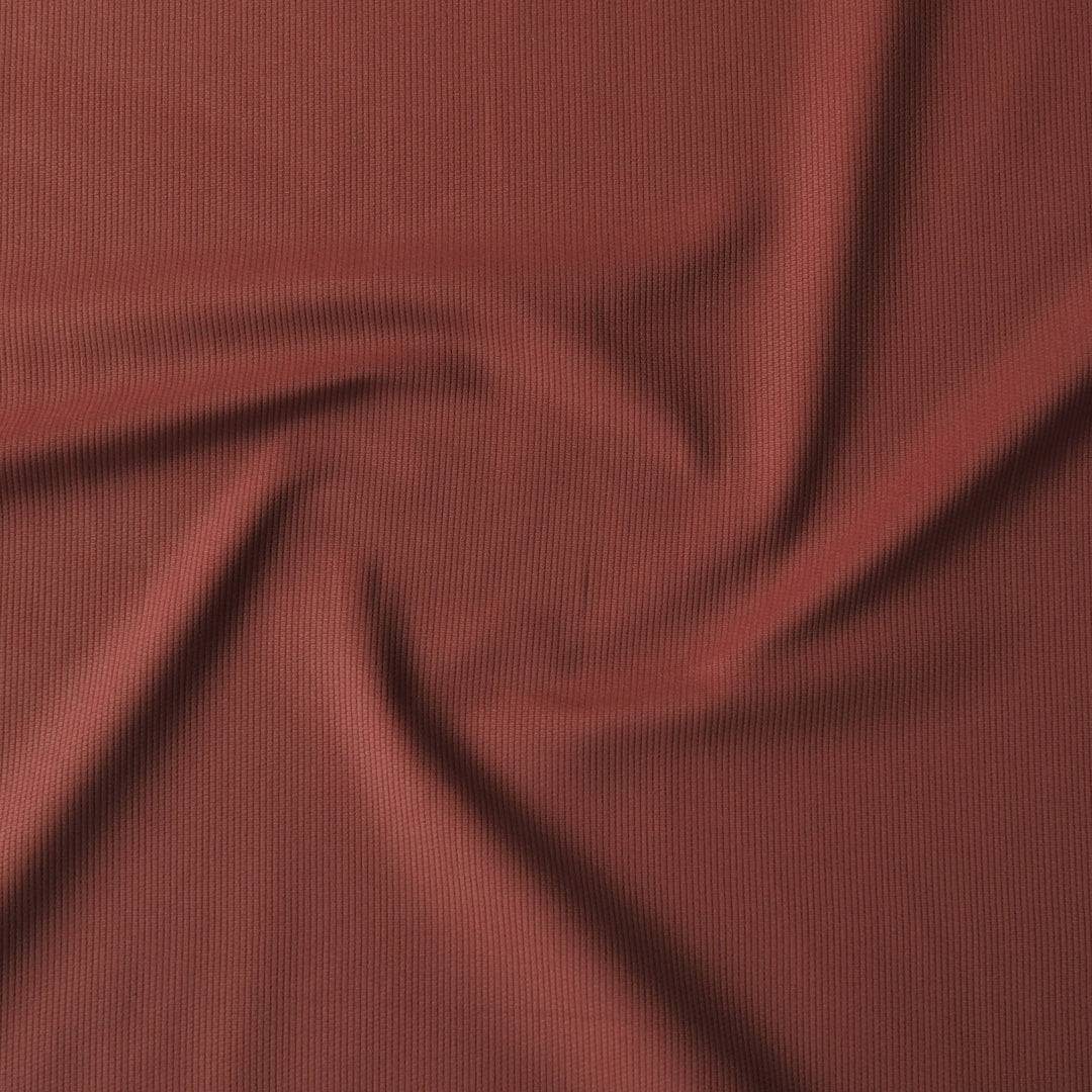 A close-up of draped ribbed fabric in a muted brick red or terracotta color, filling the entire frame.