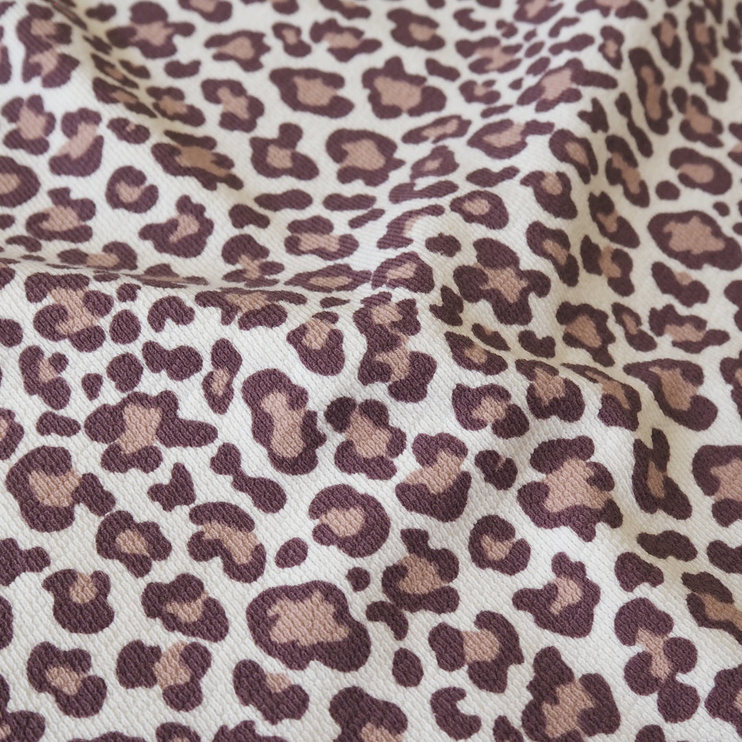 A close-up of leopard print fabric with a cream background and dark brown and tan spots, showing a soft woven texture with gentle folds.