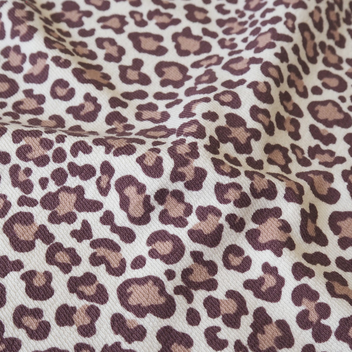 A close-up of leopard print fabric with a cream background and dark brown and tan spots, showing a soft woven texture with gentle folds.