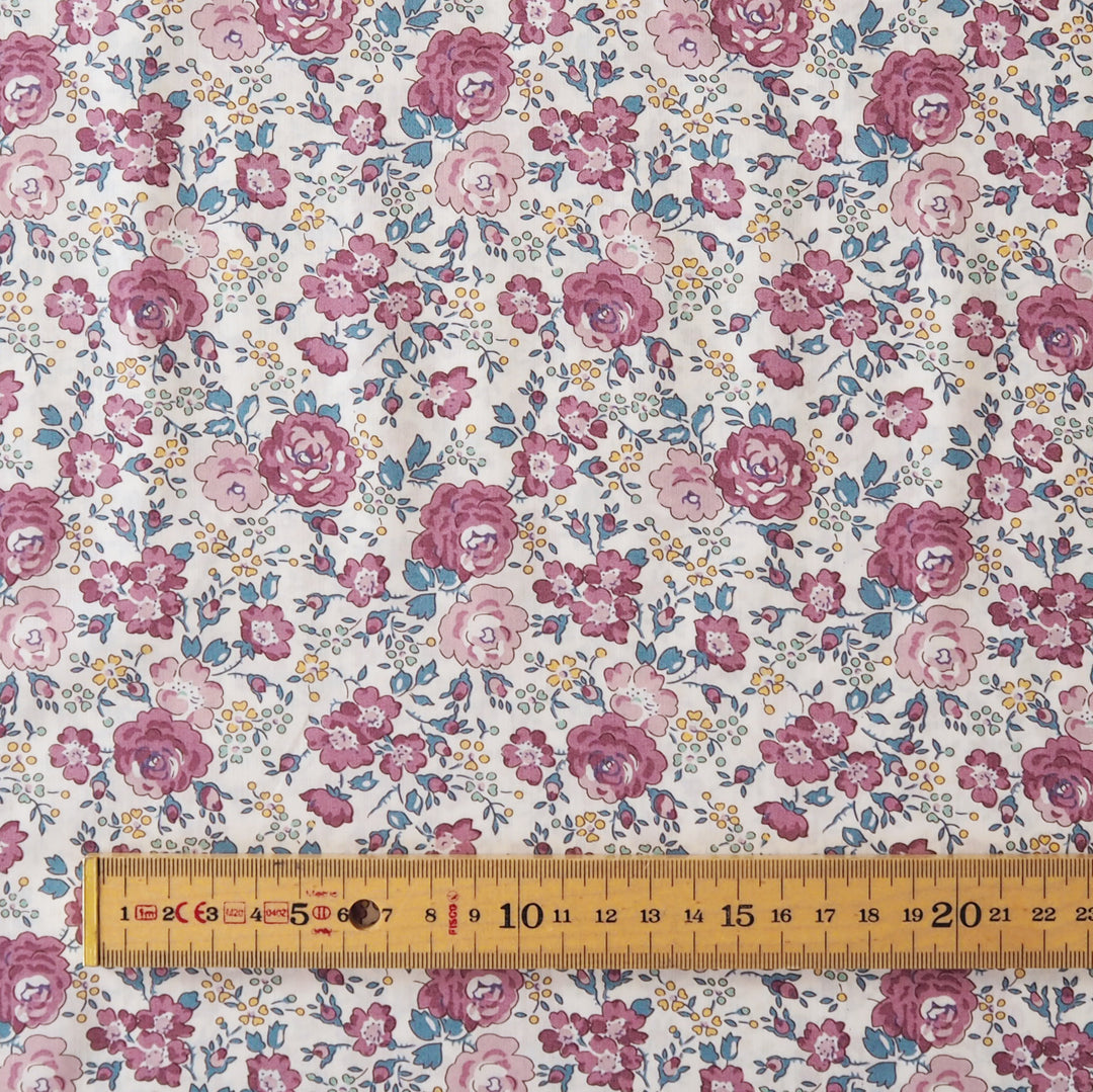 A close-up of a floral fabric with a repeating pattern of pink and mauve roses, blue leaves, and small yellow flowers on a white background, with a wooden ruler along the bottom edge for scale.