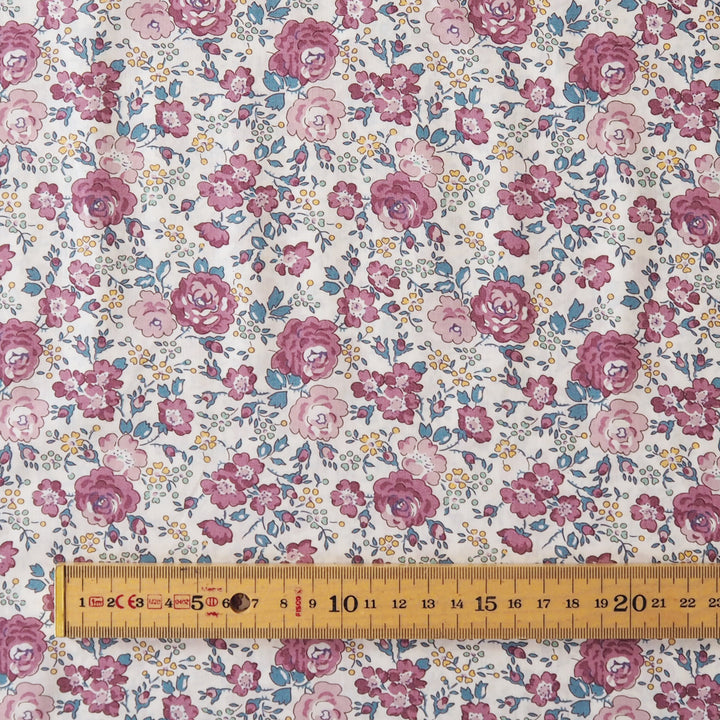 A close-up of a floral fabric with a repeating pattern of pink and mauve roses, blue leaves, and small yellow flowers on a white background, with a wooden ruler along the bottom edge for scale.