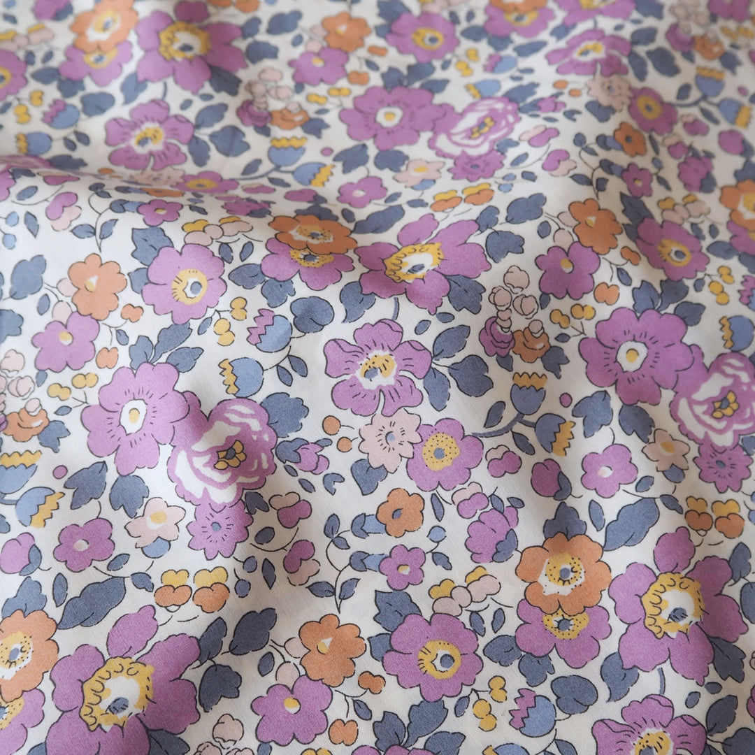 A close-up of a floral fabric with a dense pattern of purple, orange, and blue flowers on a cream background.