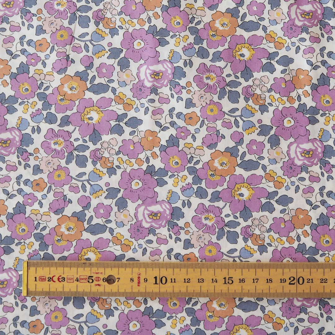 A close-up of a floral fabric pattern with purple, pink, orange, and blue flowers on a white background, with a wooden ruler along the bottom edge showing measurements.