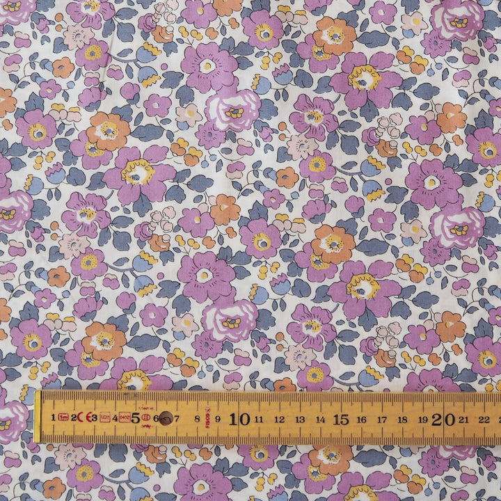 A close-up of a floral fabric pattern with purple, pink, orange, and blue flowers on a white background, with a wooden ruler along the bottom edge showing measurements.