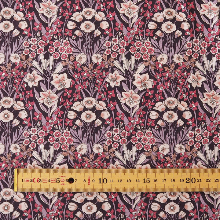 A close-up of a floral fabric pattern with pink and cream flowers, green stems, and small red berries on a dark purple background, with a yellow wooden ruler along the bottom edge for scale.