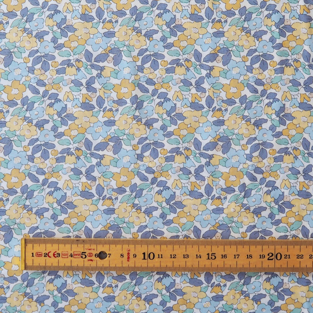 A close-up of a fabric or wallpaper pattern featuring small yellow and white flowers with blue and purple leaves on a white background, with a wooden ruler at the bottom showing the scale.