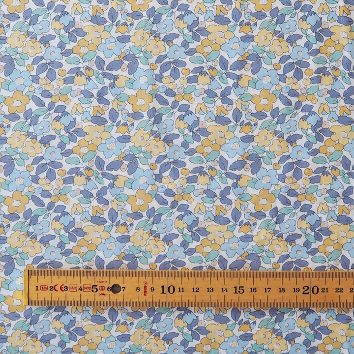 A close-up of a fabric or wallpaper pattern featuring small yellow and white flowers with blue and purple leaves on a white background, with a wooden ruler at the bottom showing the scale.