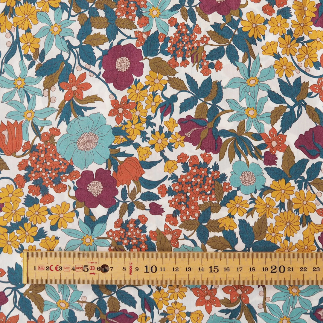A close-up of a floral fabric pattern with large flowers in teal, magenta, yellow, orange, and brown on a white background, with a wooden ruler along the bottom edge showing scale.