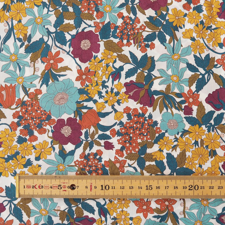 A close-up of a floral fabric pattern with large flowers in teal, magenta, yellow, orange, and brown on a white background, with a wooden ruler along the bottom edge showing scale.
