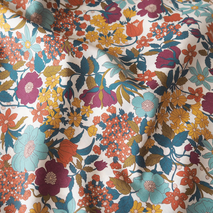 A close-up of a floral fabric with a dense, multicolor pattern featuring teal, magenta, orange, yellow, and olive green flowers and leaves on a cream background.