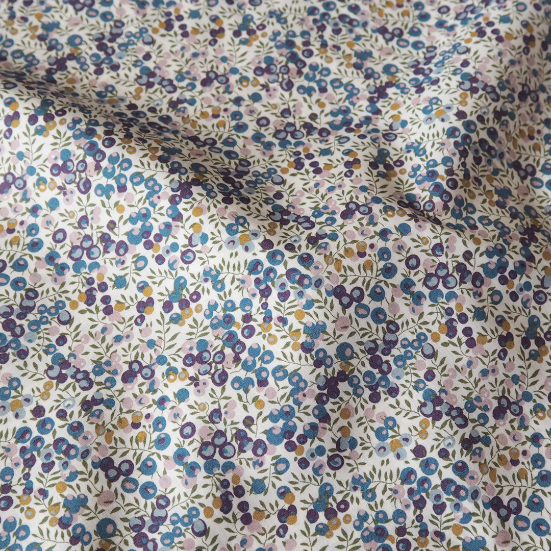 A close-up of a fabric surface with a dense small floral print on a white base. The pattern includes blue, purple, pink, and yellow flowers with green stems and leaves, arranged in a scattered all-over print.