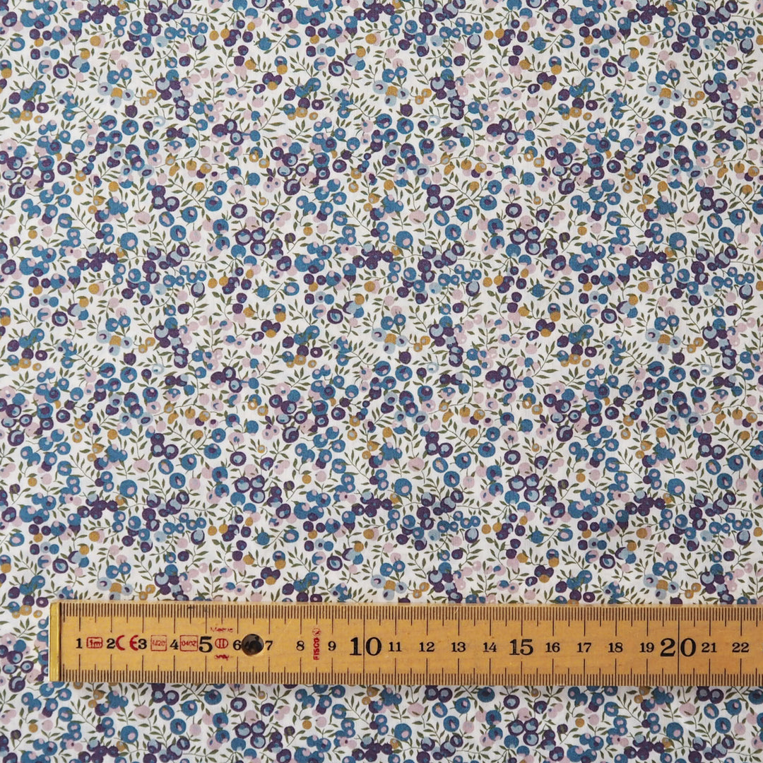 A close-up of a floral fabric pattern with small blue, purple, yellow, and green flowers on a white background, with a wooden ruler along the bottom edge for scale.