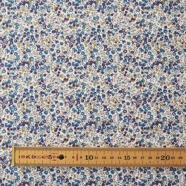 A close-up of a floral fabric pattern with small blue, purple, yellow, and green flowers on a white background, with a wooden ruler along the bottom edge for scale.