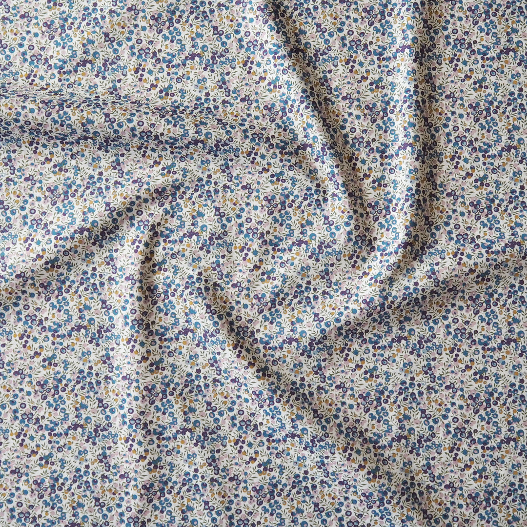 A close-up of a draped fabric with a dense, small-scale floral print in blue, purple, and yellow on a warm off-white background.