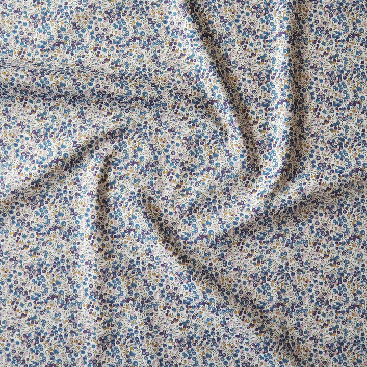 A close-up of a draped fabric with a dense, small-scale floral print in blue, purple, and yellow on a warm off-white background.